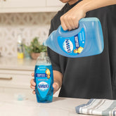 Person pouring Dawn dish soap from a large container into a smaller bottle on a kitchen counter.